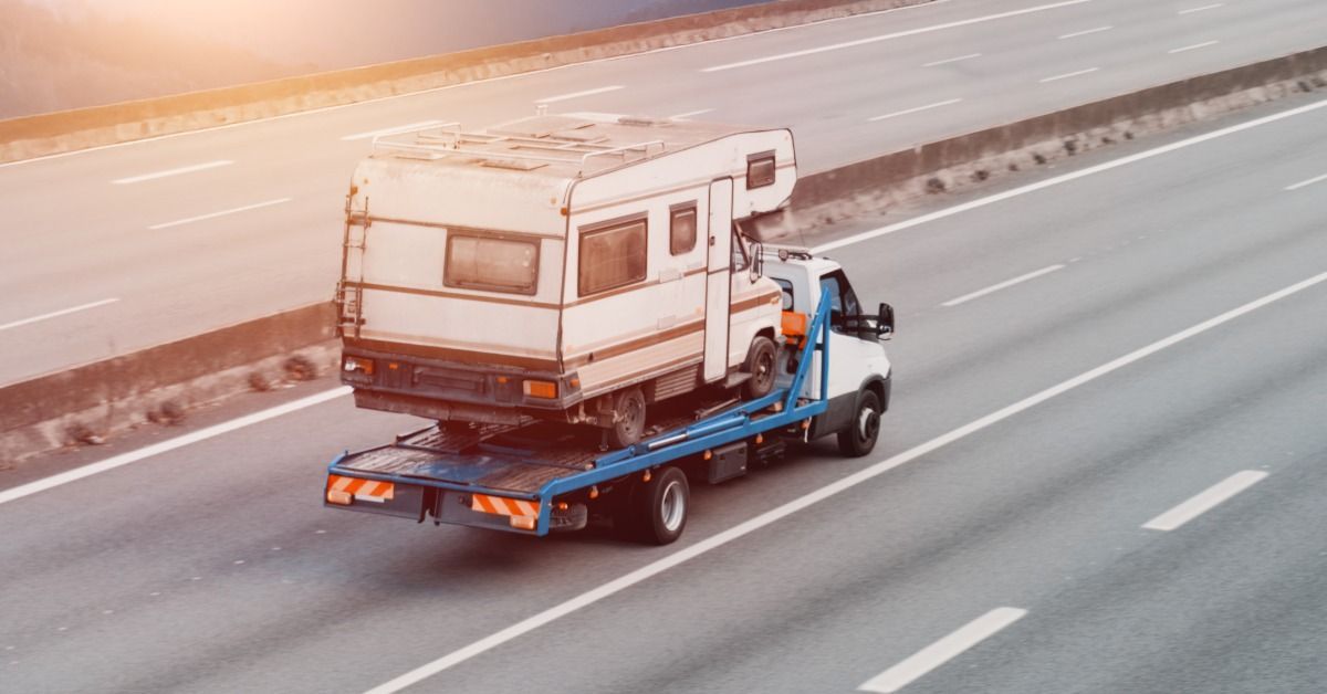 A tow truck carrying a vintage camper on a paved road, with green grass and trees visible behind it.