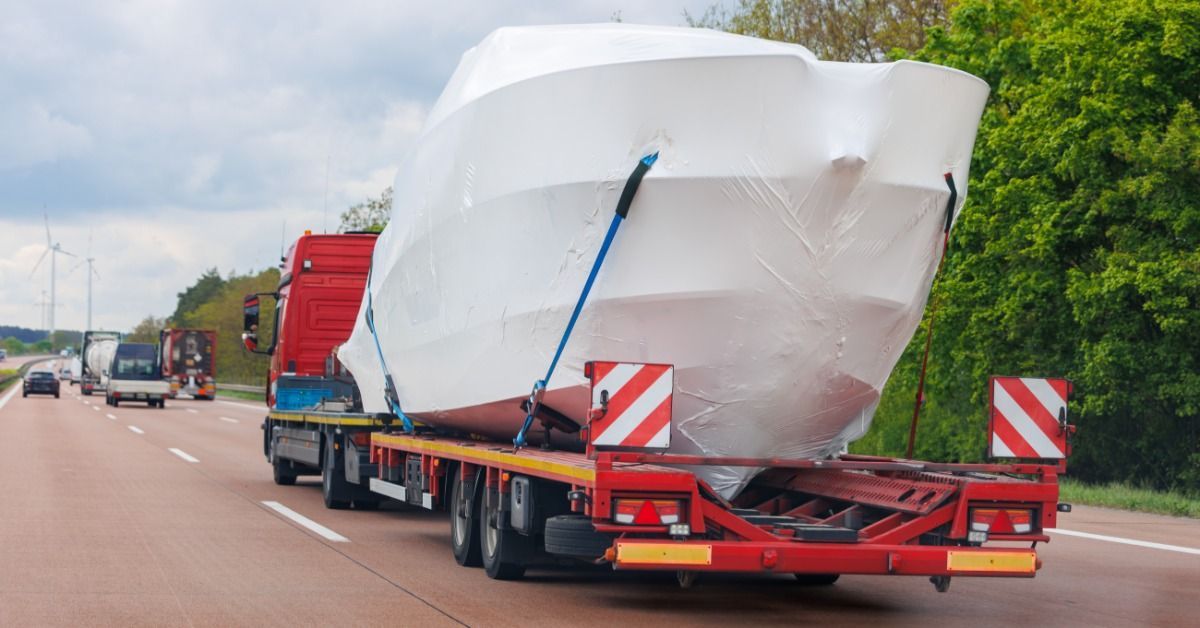 A large boat wrapped in white shrink wrap is strapped to the back of a long bed trailer on a semi on