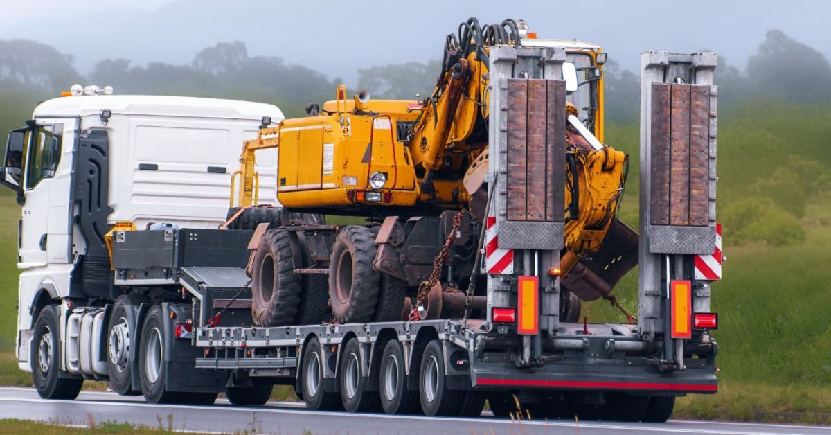 A semi carries a large piece of heavy construction equipment on a flatbed trailer. The road pavement