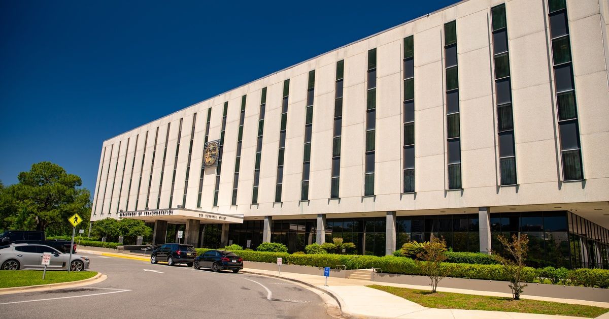 A beige building labeled “Department of Transportation” features a glass entrance, parked cars, and trimmed greenery.