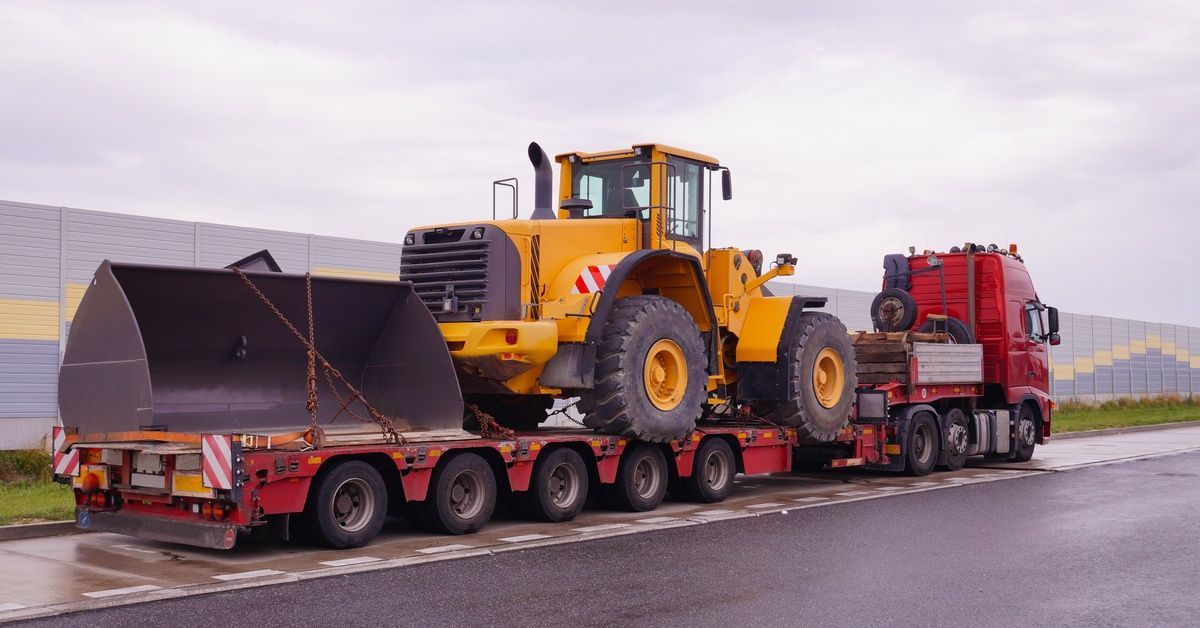 A red semi-trailer parked on a road shoulder carries a bulldozer, with a gray and yellow fence and cloudy sky in view.