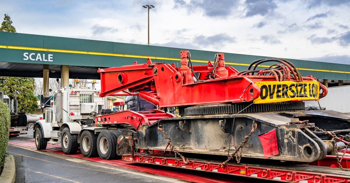 A white semi-truck with a step-down trailer hauls red equipment marked “Oversize Load” while entering a scale station.