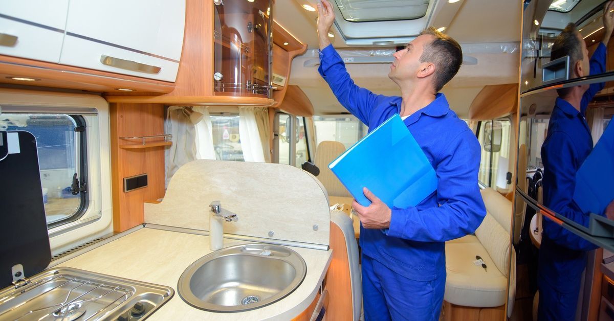 A man in blue coveralls examining the ceiling of a camper van while standing near a countertop with a sink and stovetop.