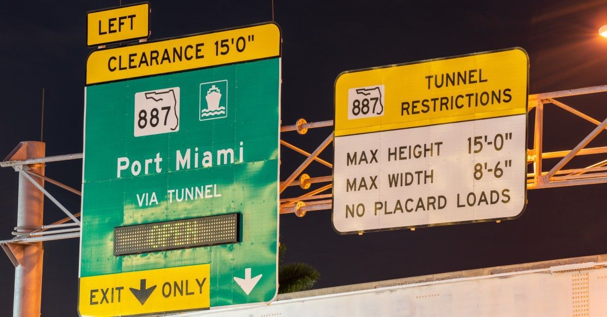 Two gantry signs at night. One labels the exit for Port Miami, and the other lists vehicle dimension restrictions for the tunnel.