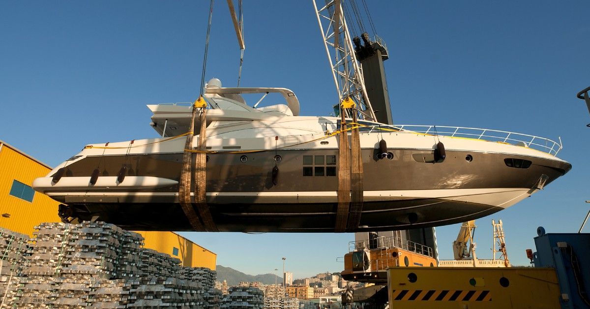 A crane lifts a white and gray yacht over stacked materials under clear blue skies during the daytime.