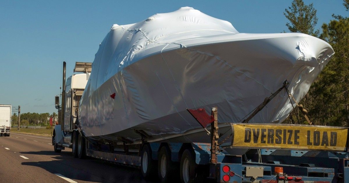A truck carries a boat covered by a white tarp with an “OVERSIZE LOAD” banner on the back.
