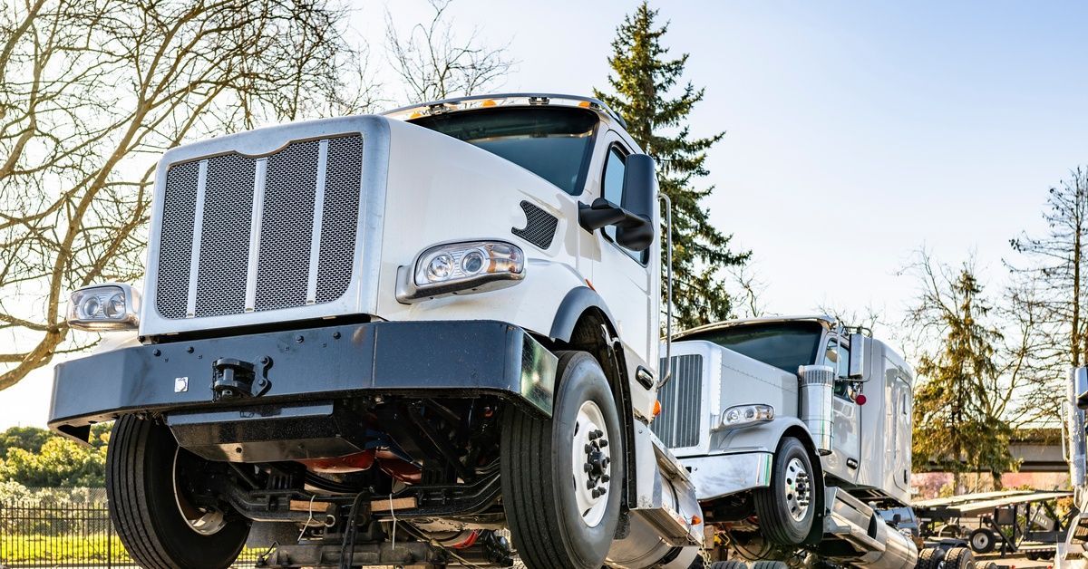 Two big white rigs are attached by their front axles to a tractor frame. Leafless trees are behind them in soft light.