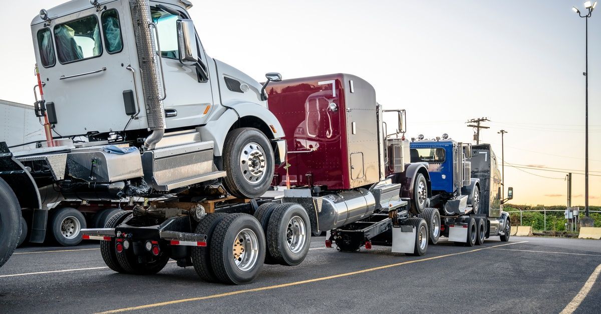 A charcoal gray semi-truck forms a road train with three big rigs stacked in a lot under a setting s