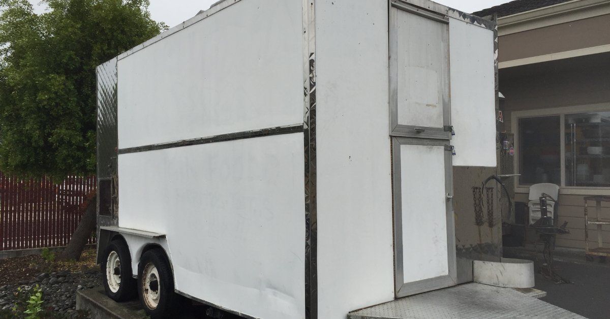 A white concession trailer parked on concrete beside a brown building, with a fence and a tree behind the trailer.