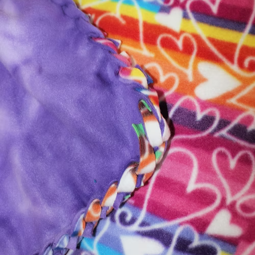 Purple tie-dye fleece blanket next to a blanket with pink, red, and orange hearts; edges are knotted together.