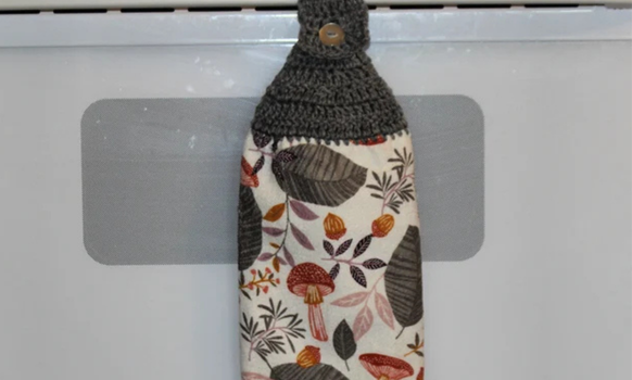 Crocheted kitchen towel hanging on a stove handle with a mushroom and leaf pattern on the fabric.