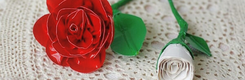 Close-up of a red paper rose and a closed white paper rose with green stem on a lacy surface.