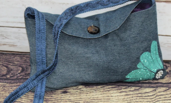 Denim purse with flap closure, decorative flower, and strap on a wooden surface.