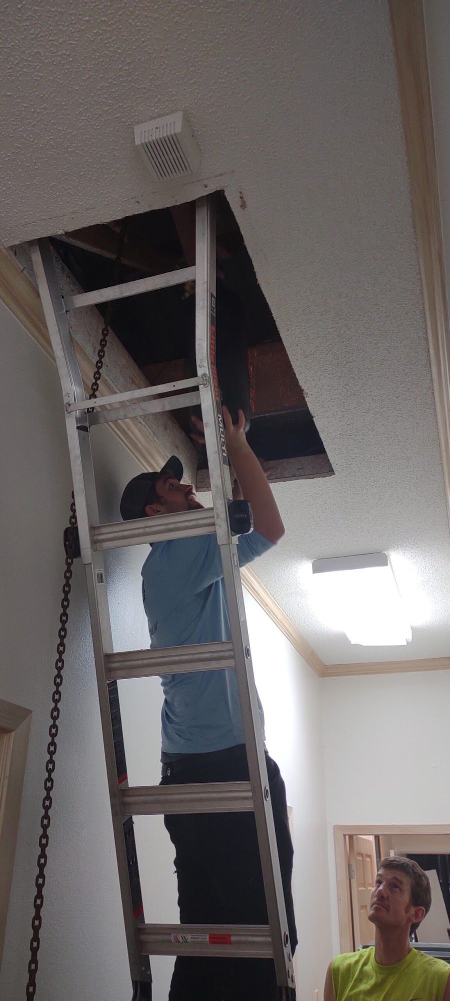 A person climbs a ladder to inspect an open attic hatch while another person watches from below.