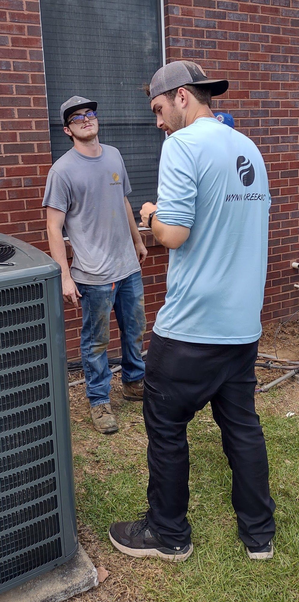 Two people in hats and casual clothing stand outside near a brick wall and an outdoor HVAC unit.