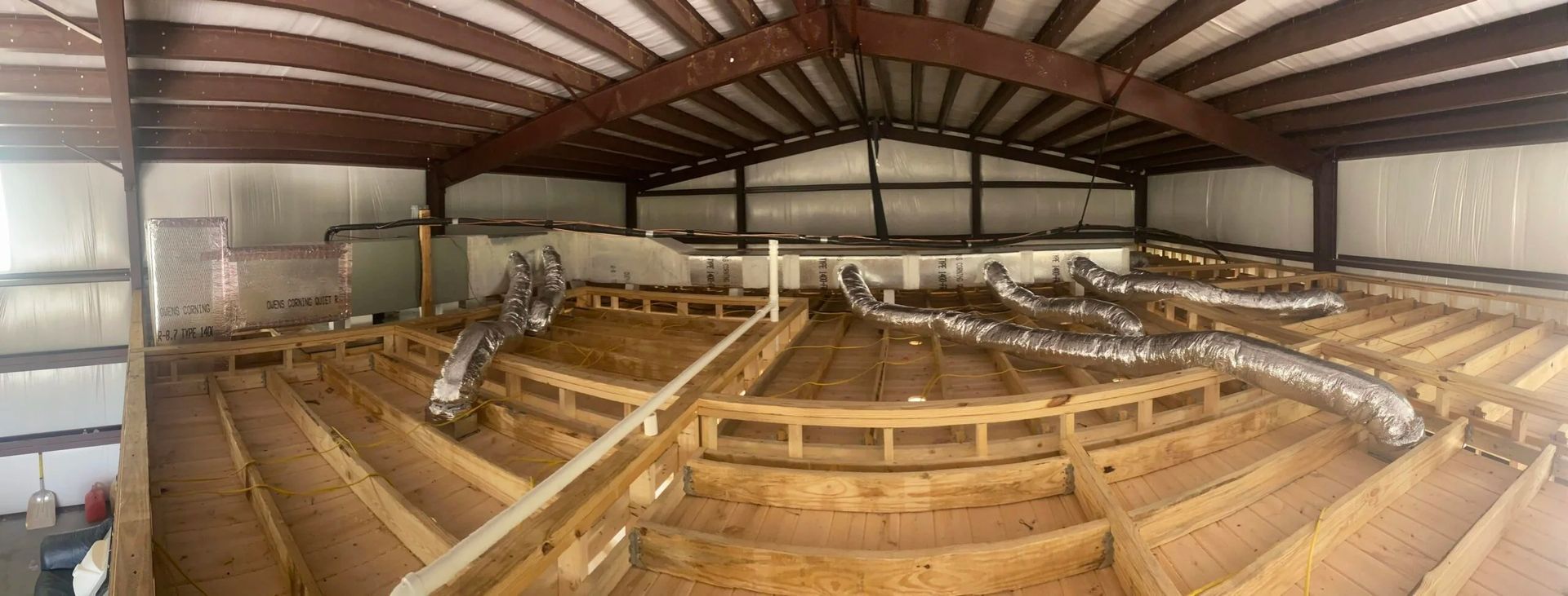 A panoramic view of an unfinished attic space with exposed wooden floor joists, flexible air ducts, and metal roof trusses.