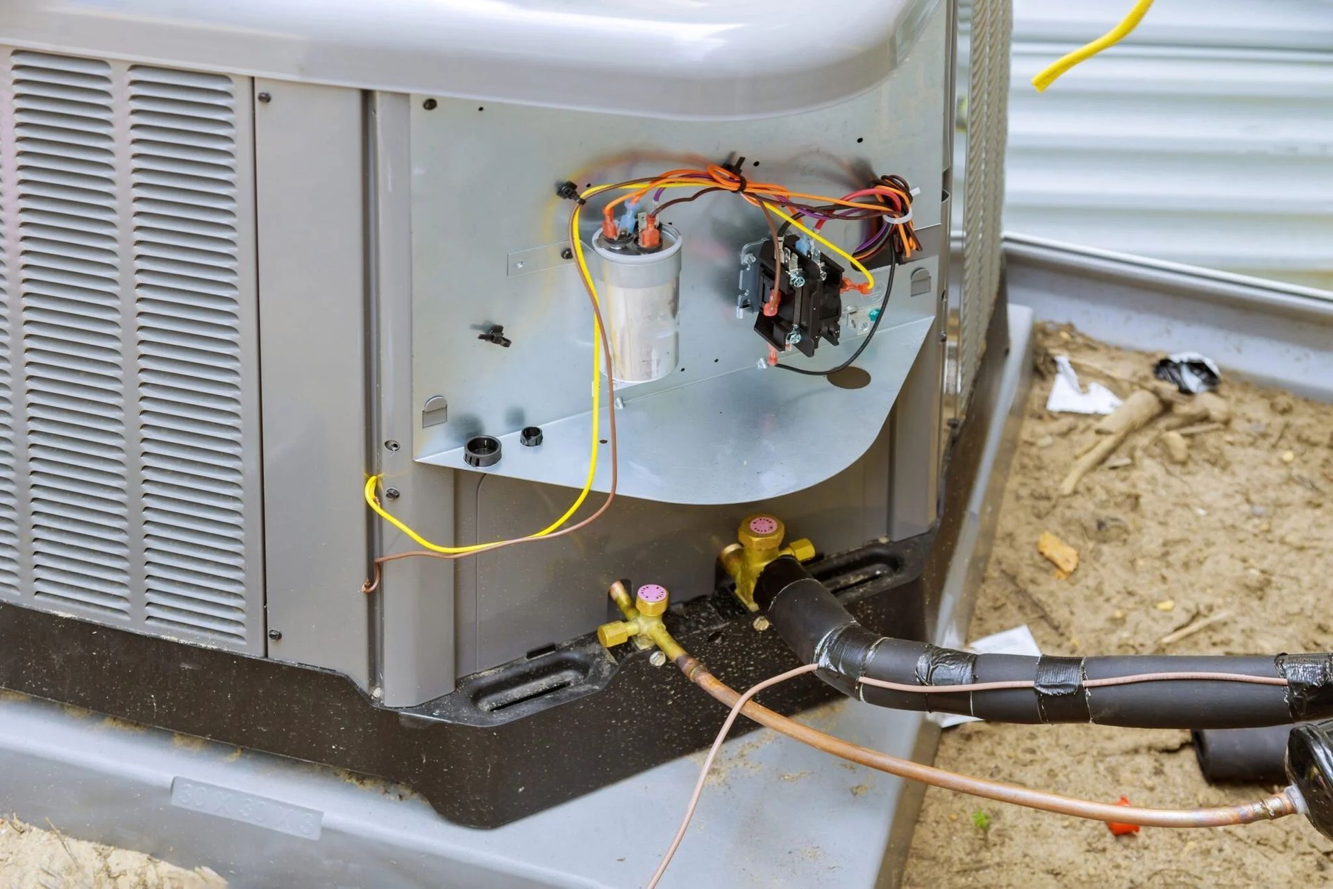 An outdoor HVAC condenser unit with its electrical panel open, revealing wiring and a capacitor above refrigeration lines.