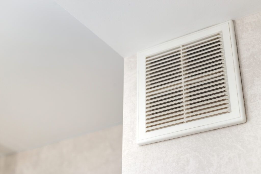 A white, square air vent grille mounted on a light-colored, textured wall in a room.