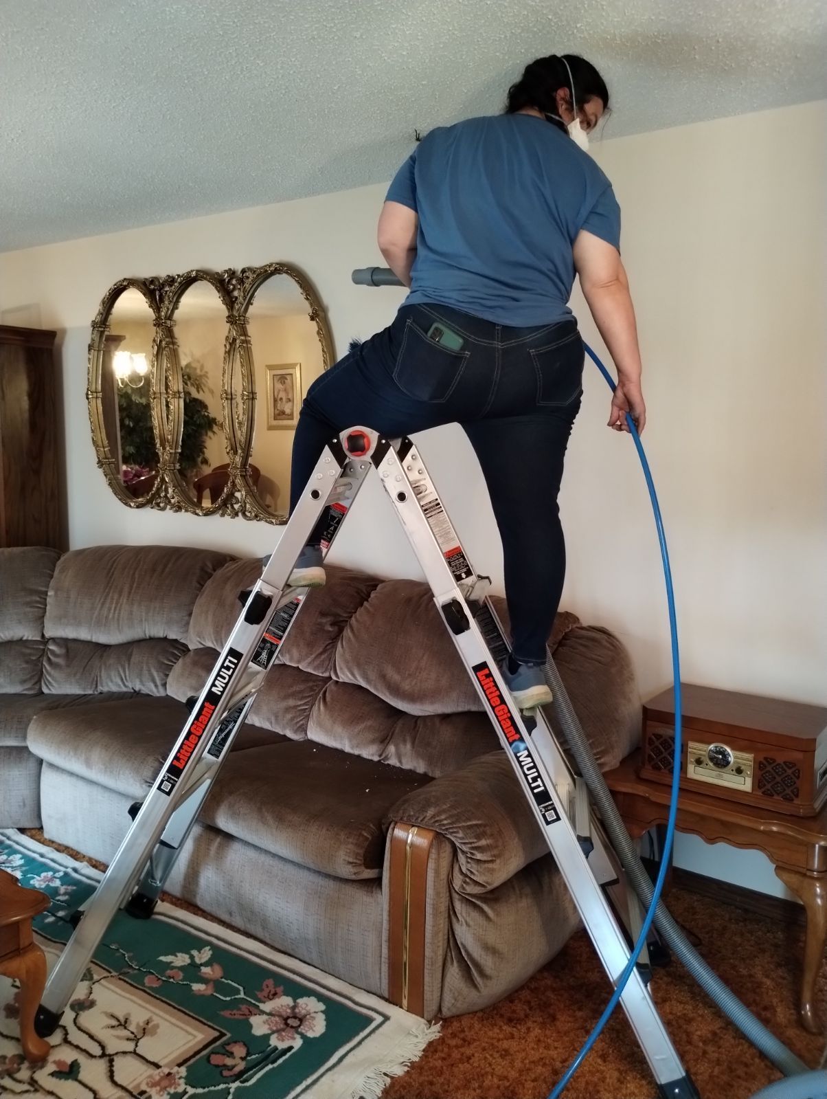 A person stands on a folding ladder in a living room, holding a blue hose up to clean a patch on the ceiling.