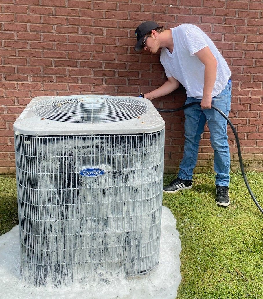 A person sprays cleaning foam on an outdoor HVAC unit next to a brick wall.