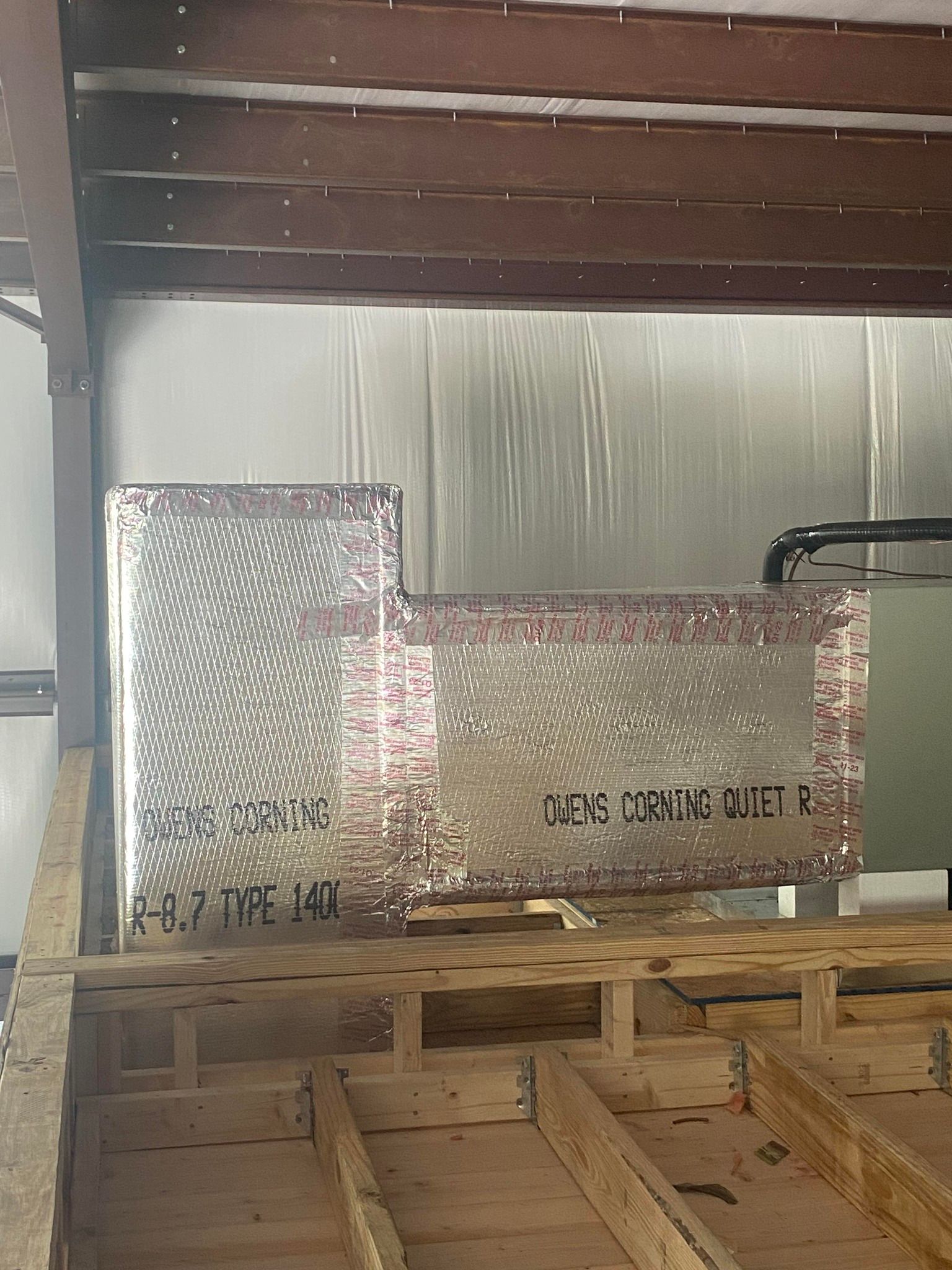 Rectangular HVAC ductwork wrapped in silver metallic insulation, positioned on wooden floor joists in an unfinished room.