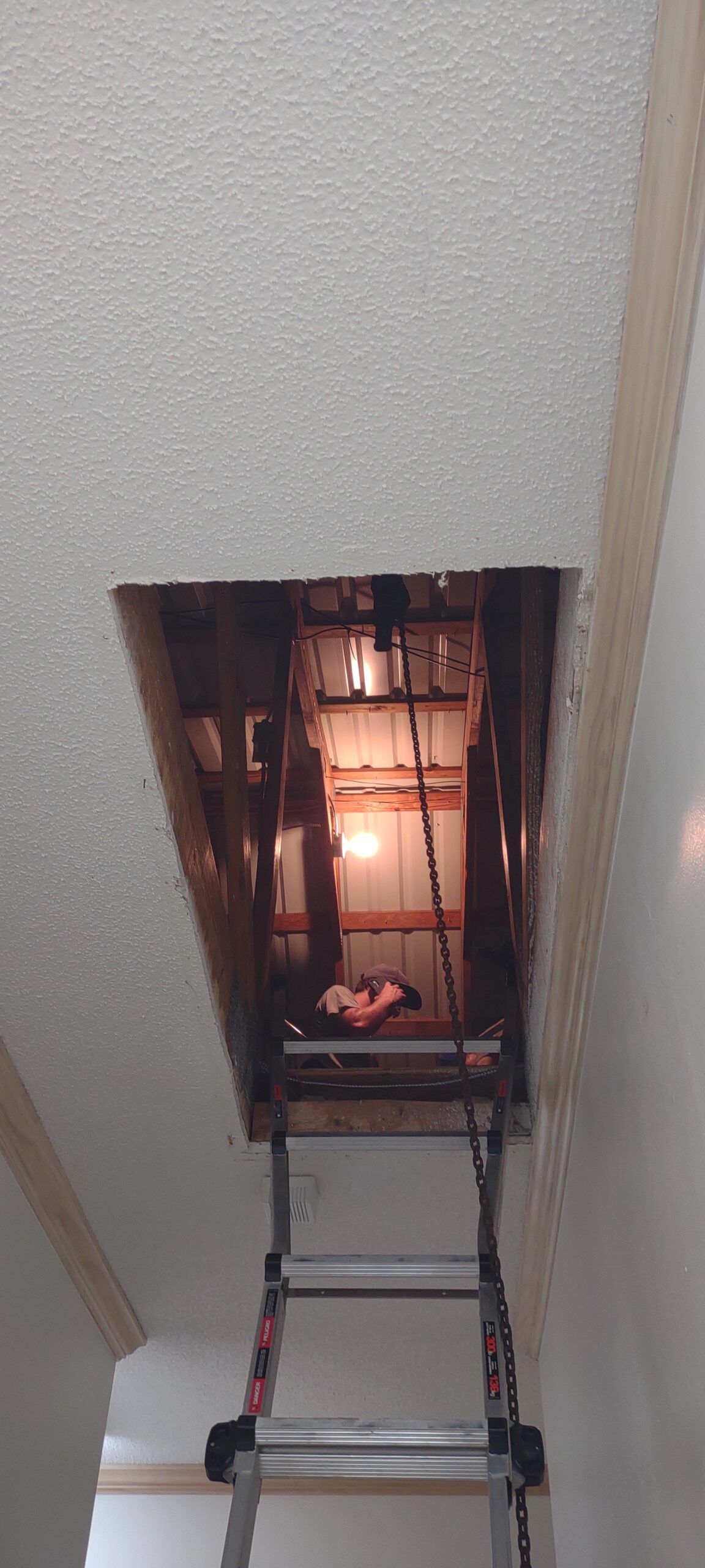 A view from a ladder looking up through a ceiling cutout into an attic space with wooden rafters and exposed wiring.