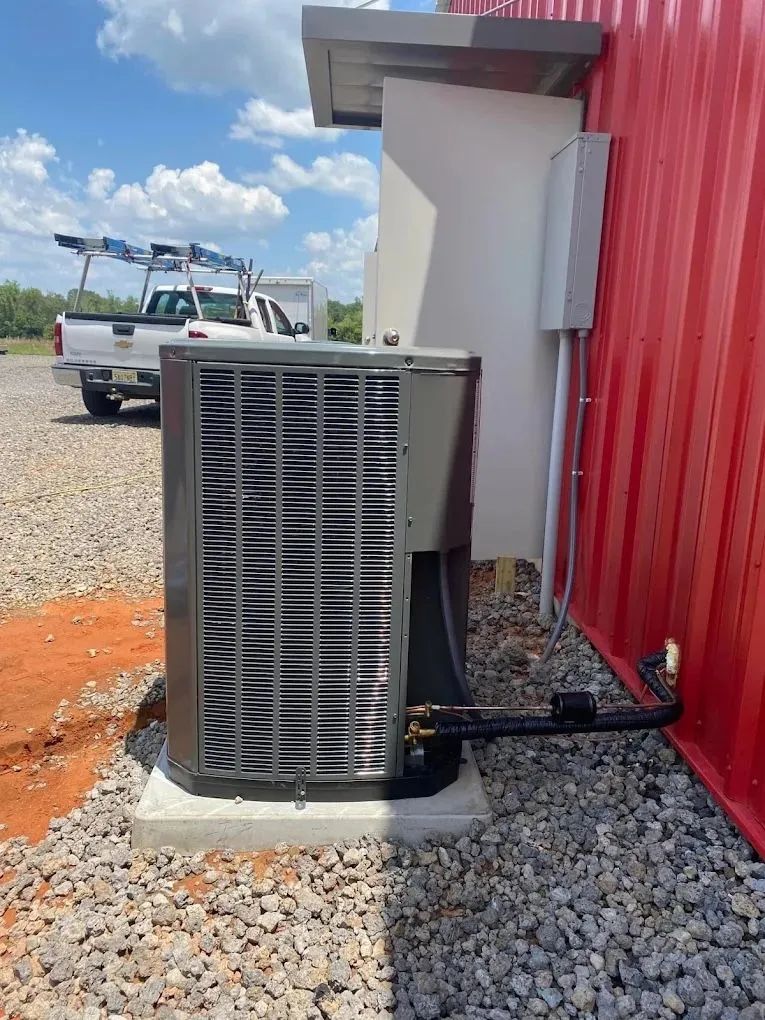 A grey outdoor HVAC condenser unit sits on a concrete pad next to a red corrugated metal building on a gravel lot.
