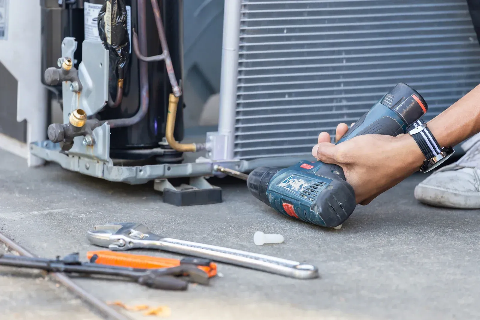 Technician using a power drill to service an outdoor HVAC unit, with tools on the ground.