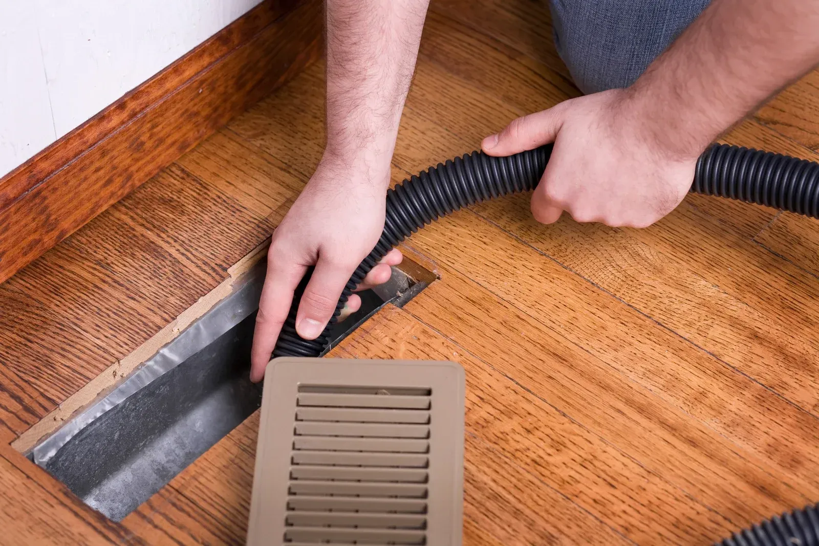 A person uses a vacuum hose to clean dust and debris from inside an open floor air vent on a hardwood floor.