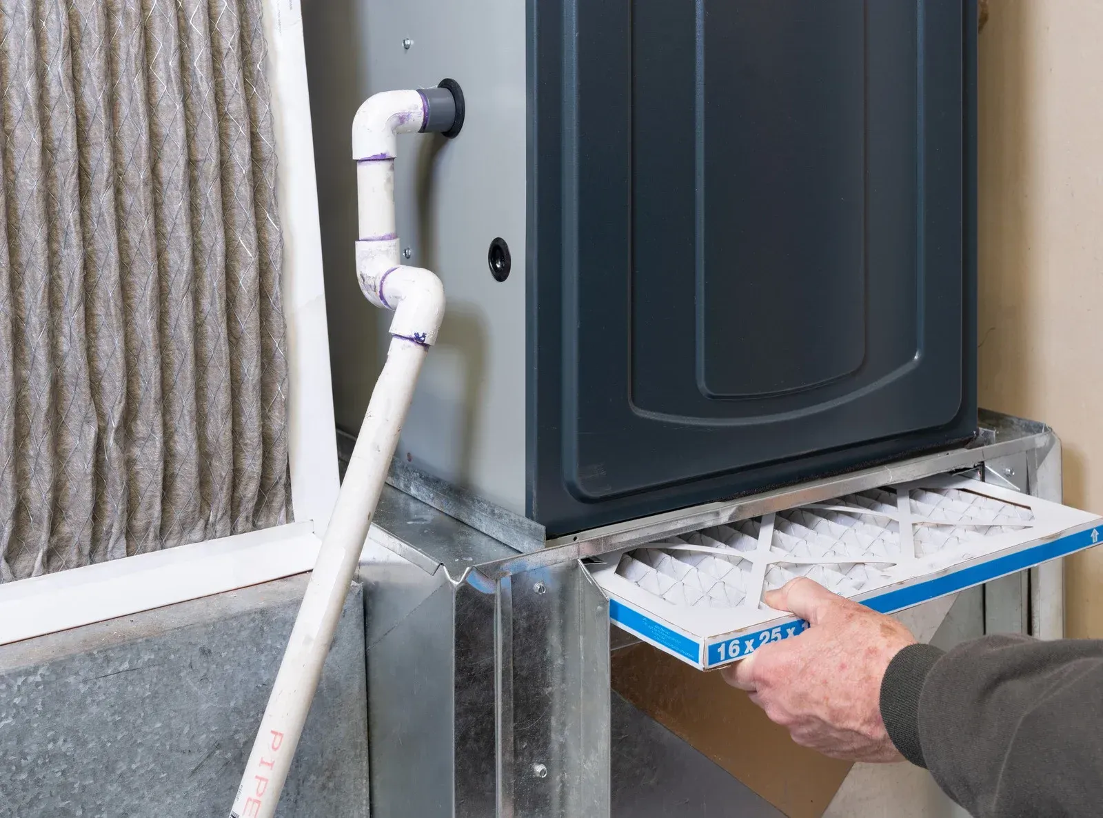 A person sliding a new pleated air filter into the bottom slot of a home furnace unit next to a dirty filter.
