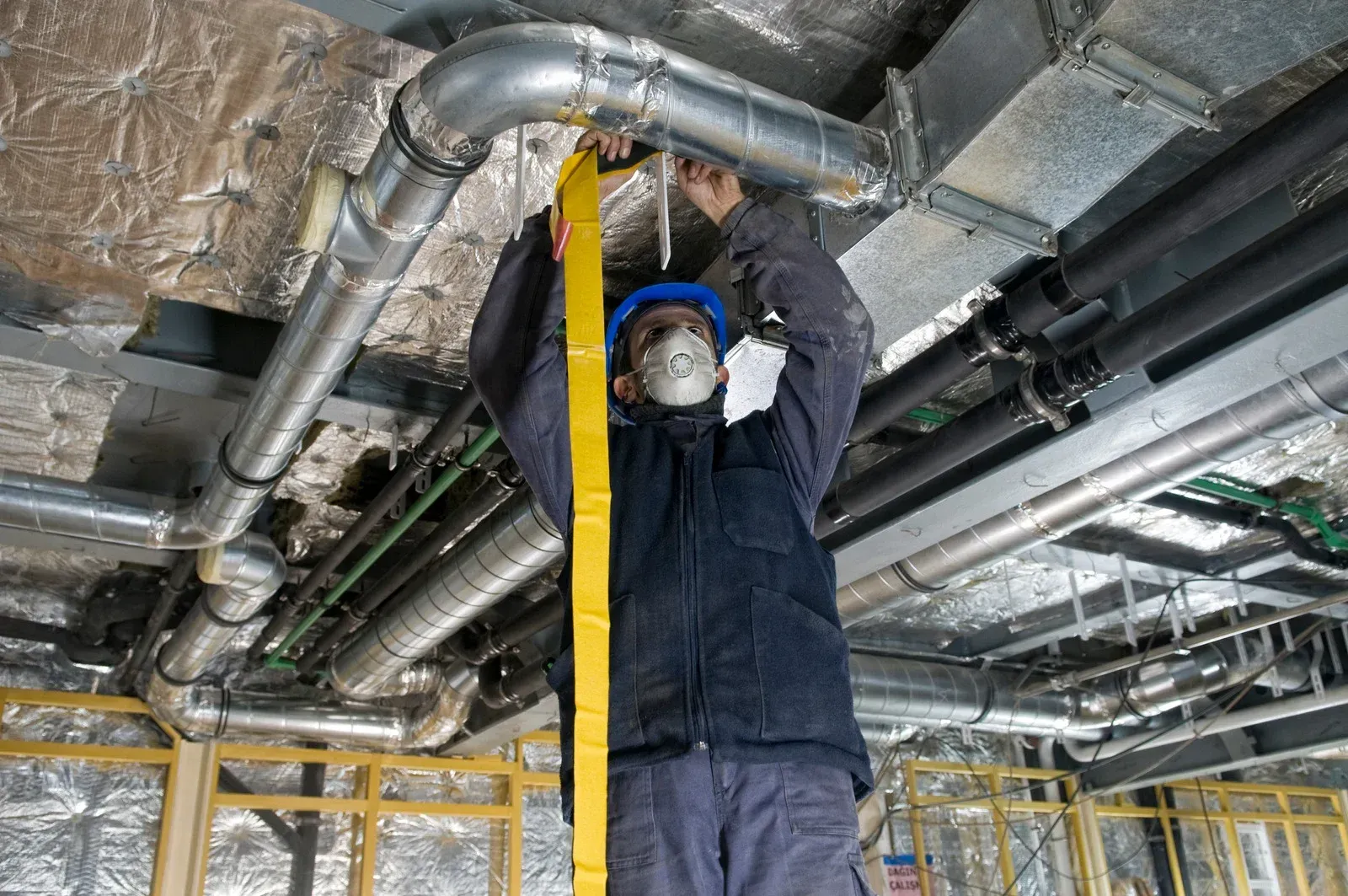 A worker wearing a hard hat and mask secures yellow tape to metal ceiling ductwork in a construction site.