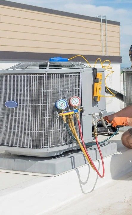 A service technician in workwear crouches on the grass to repair an outdoor air conditioning unit beside a building.