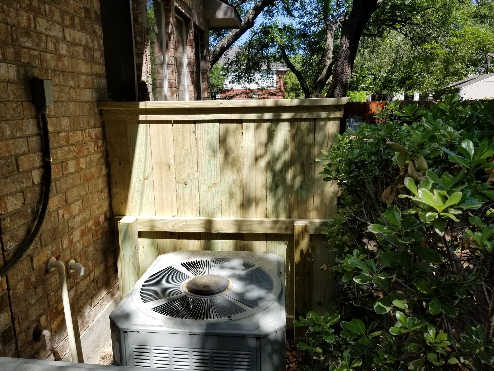 An air conditioner is sitting under a wooden fence next to a brick wall.