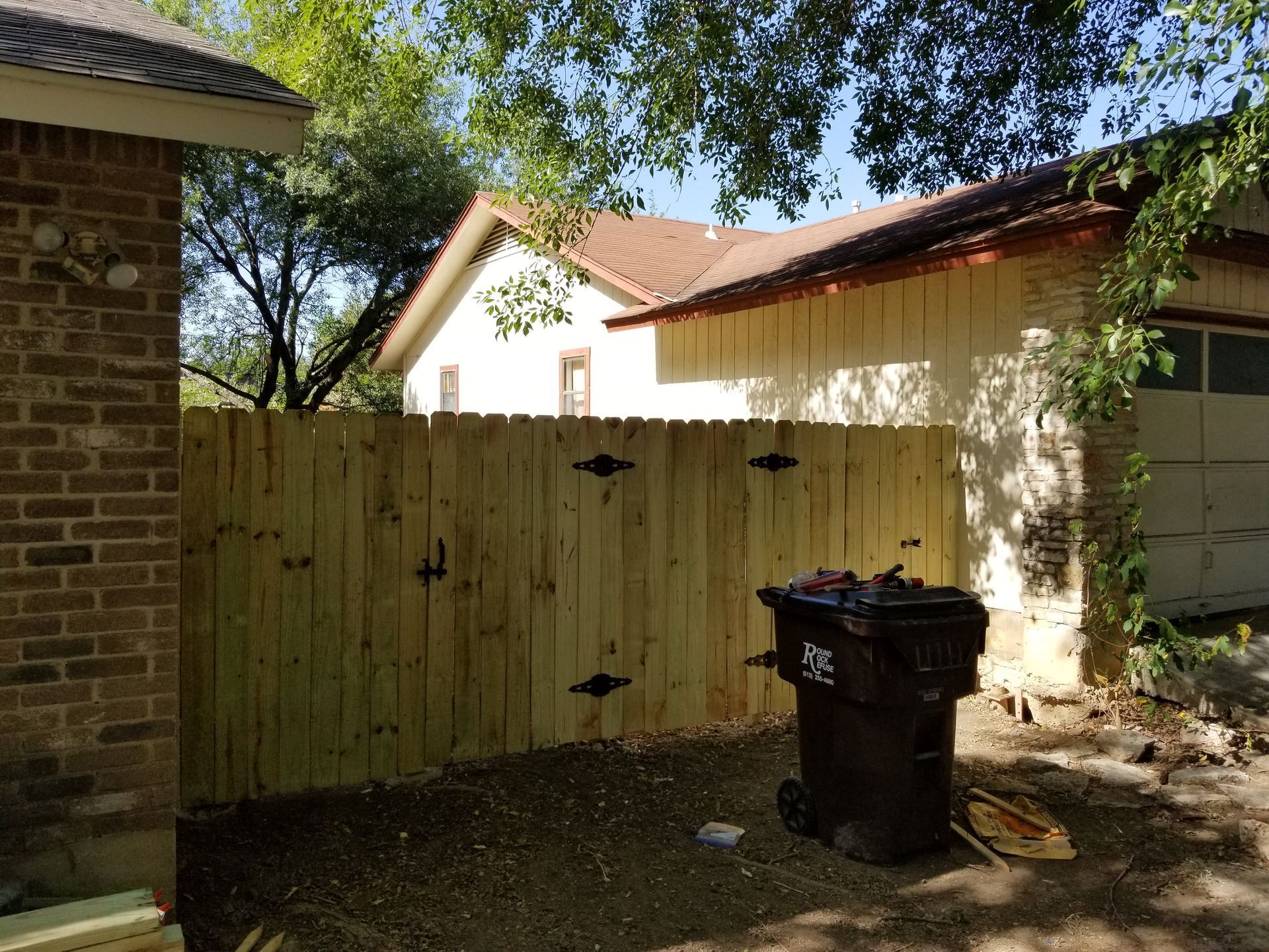 A wooden fence is surrounding a trash can in front of a brick house.