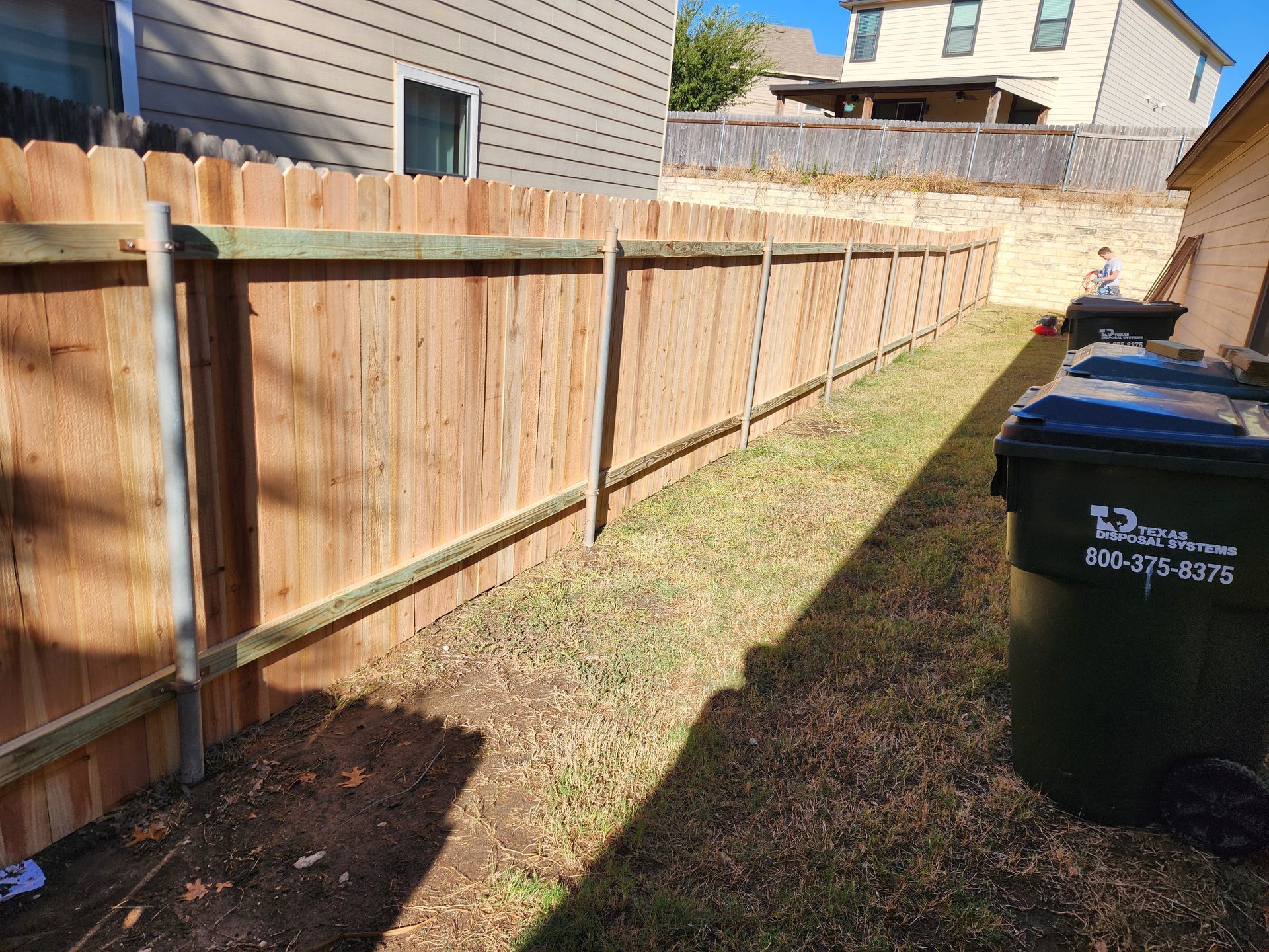 A wooden fence is surrounded by trash cans in a backyard.