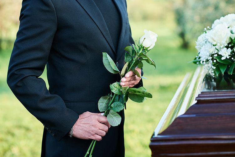 A man in a suit is holding a white rose in front of a coffin.