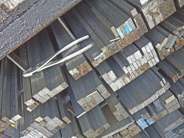 Barre di metallo di colore scuro, probabilmente acciaio, impilate in un ambiente industriale.