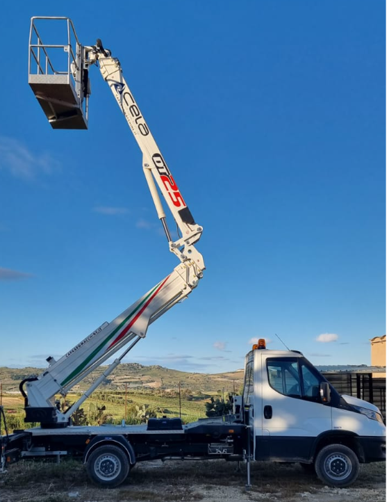 Un camion con piattaforma aerea bianca si staglia contro un cielo azzurro, con la campagna visibile sullo sfondo.