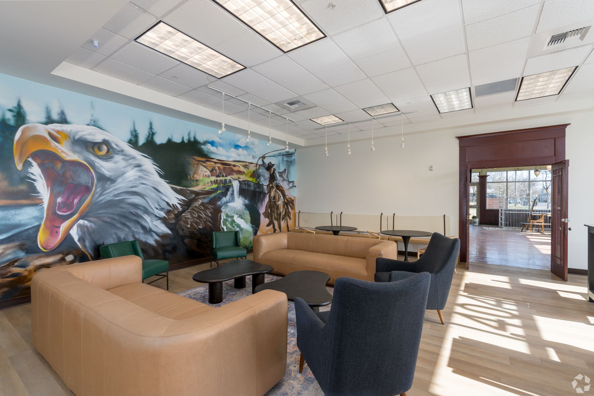 Lounge area with mural of an eagle, forest, and Native American figures. Tan and blue sofas, black tables, open doorway.
