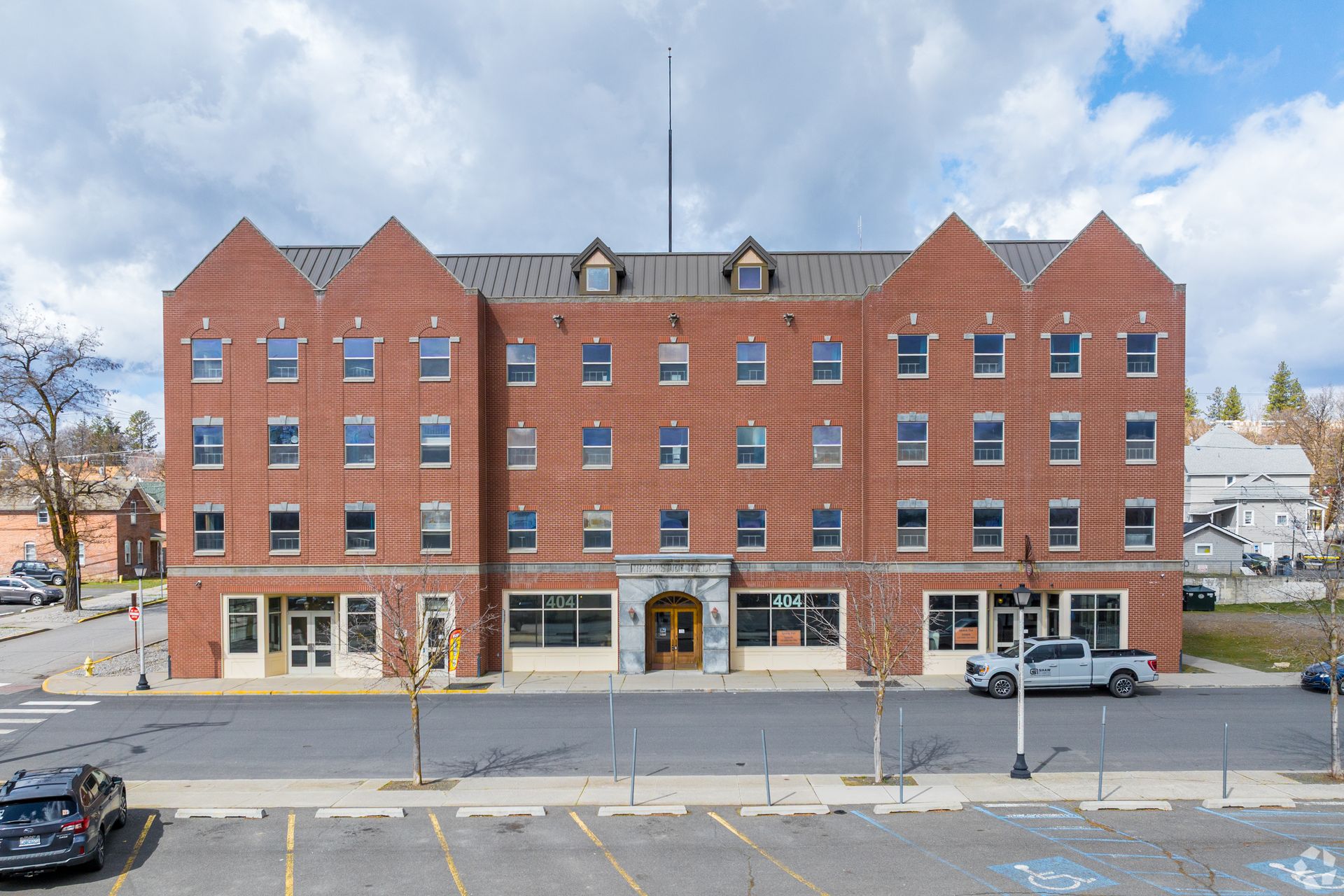 Red brick building with multiple stories and windows, a gray sky, and a parking lot in front.