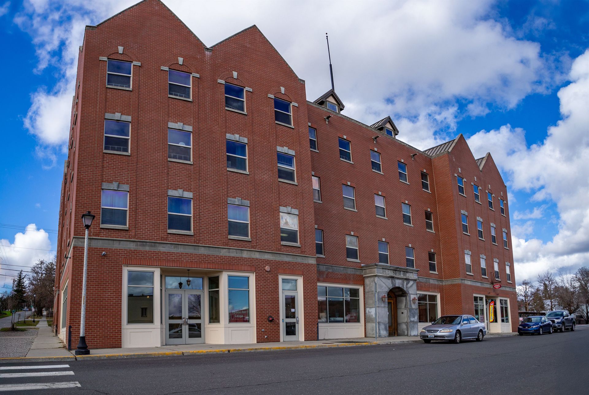 Red brick building with multiple floors and arched entrance. Cars parked in front on street.