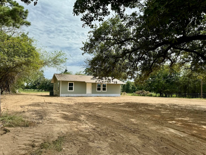 A house in the middle of a dirt field with trees in the background
