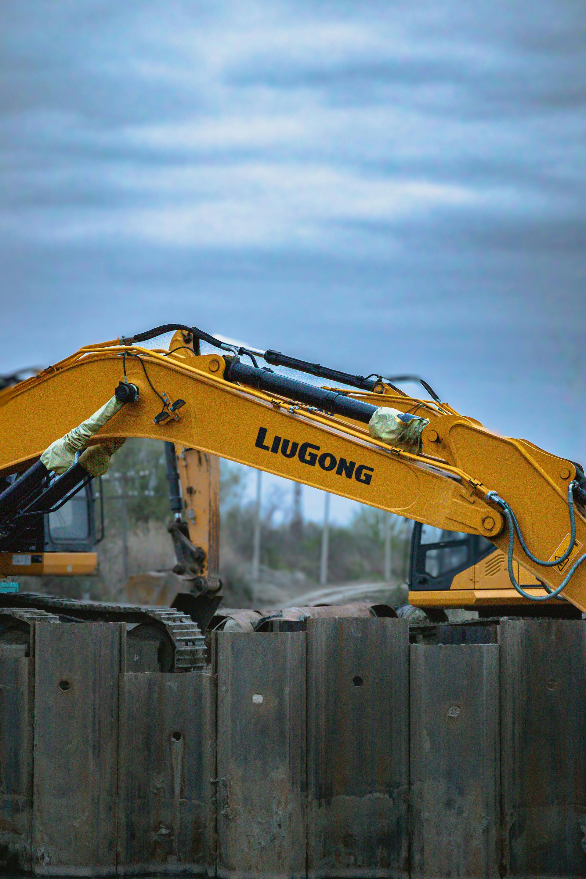 Yellow LiuGong excavator arm over a row of metal barriers at a construction site, cloudy sky.
