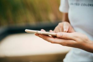 Person's hands holding a smartphone, tapping the screen. Outdoors, close-up, neutral tones.