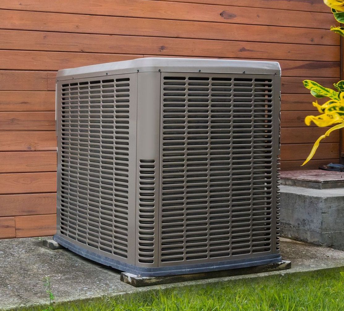 Unité de climatisation extérieure avec grilles métalliques, marron et grises, installée sur une dalle de béton près d'un mur en bois.