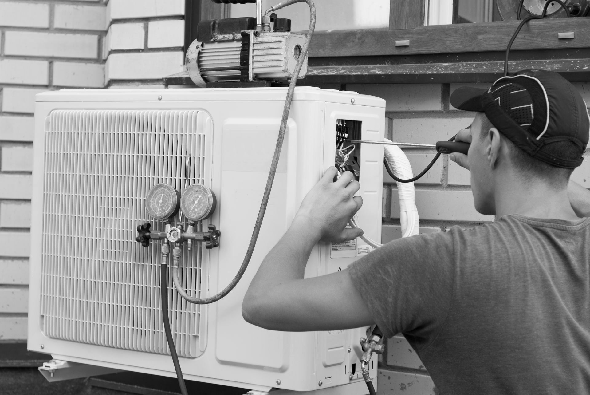 Une personne travaille sur un climatiseur extérieur, utilisant des outils et des jauges près d'un mur de briques.