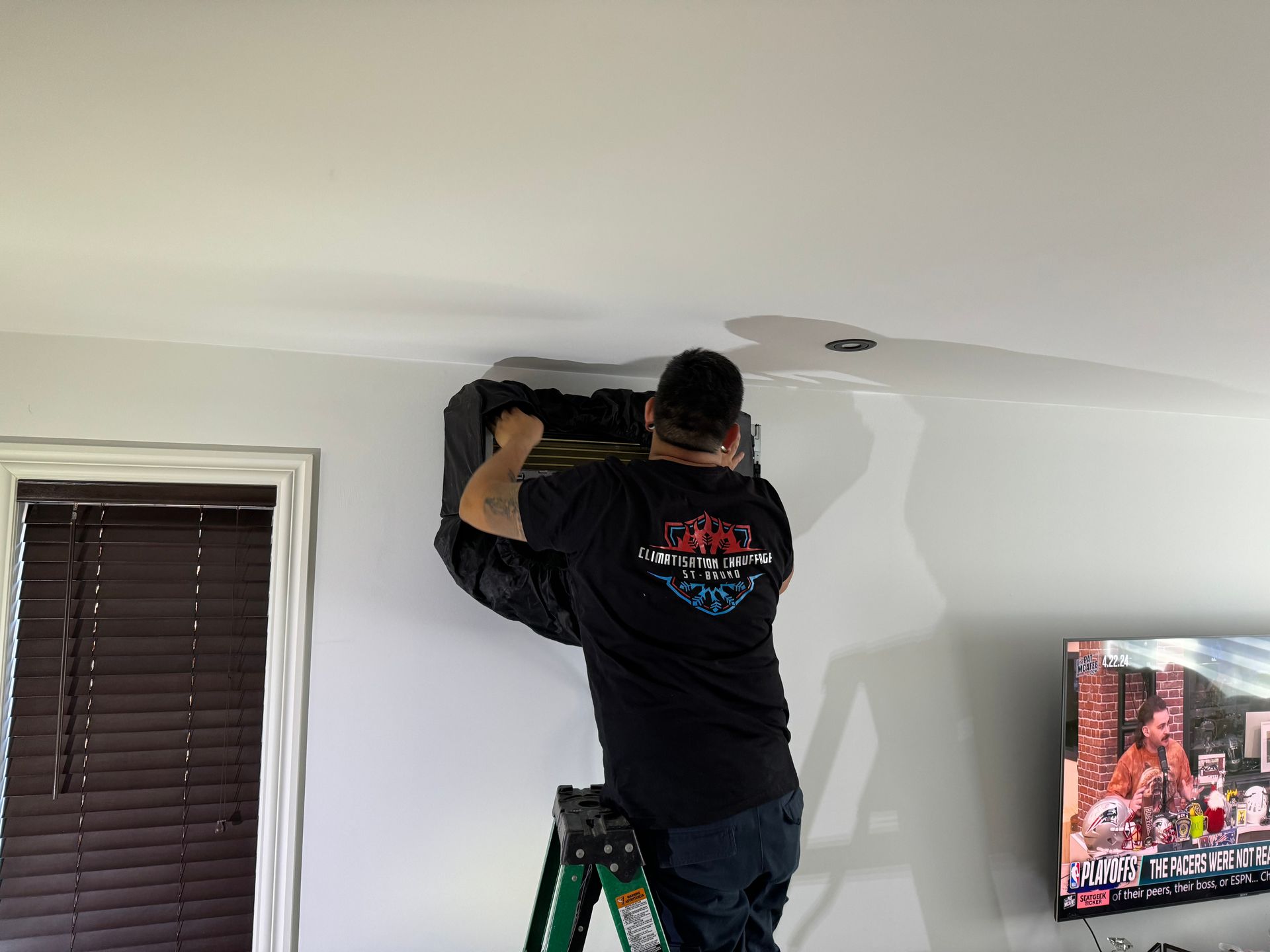 Un homme sur une échelle installe ou entretient une unité CVC dans une pièce aux murs blancs, près d'une fenêtre et d'un téléviseur.