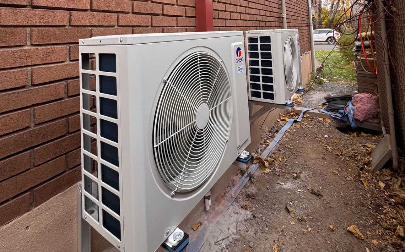 Deux unités de climatisation grises montées près d'un mur de briques rouges sur un sol humide et sale.