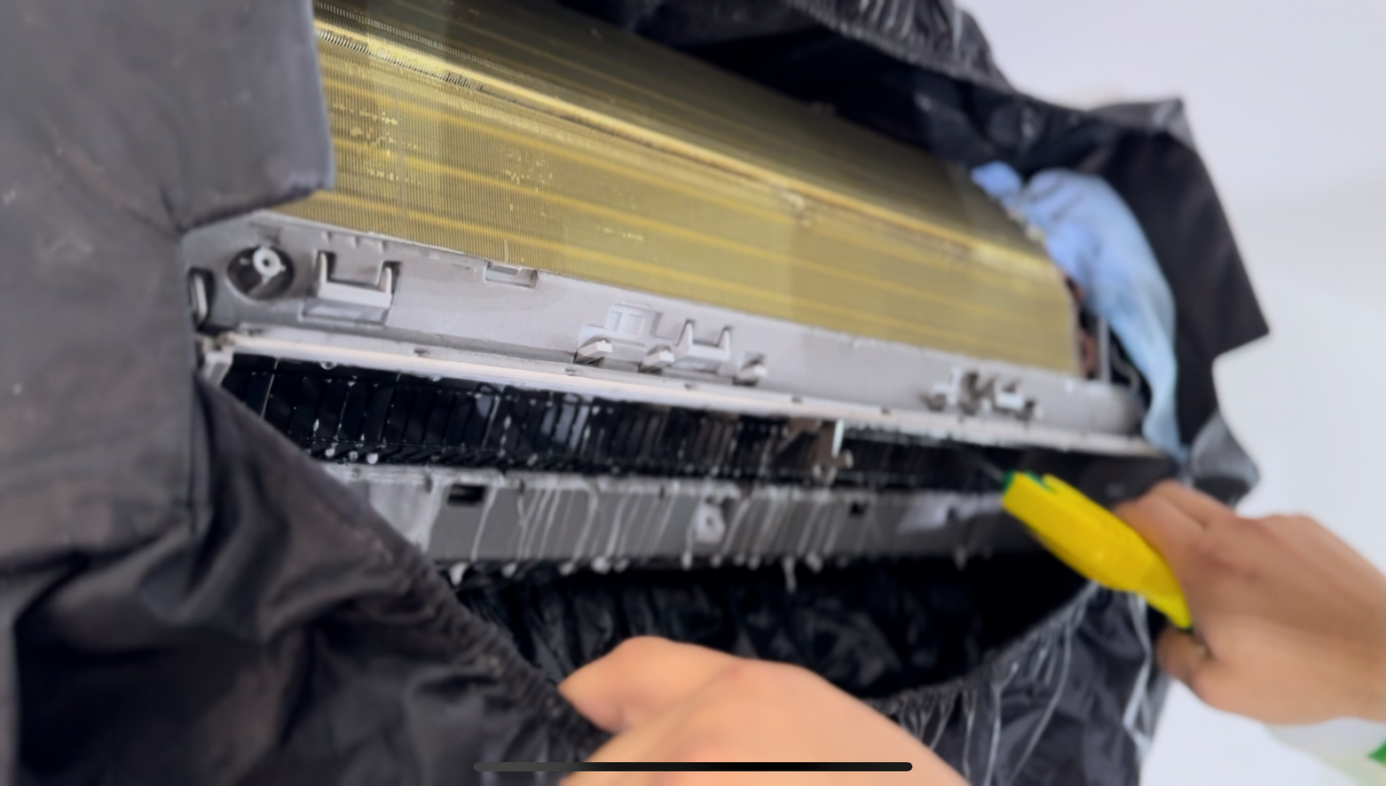 Une personne nettoie un climatiseur avec une brosse jaune.