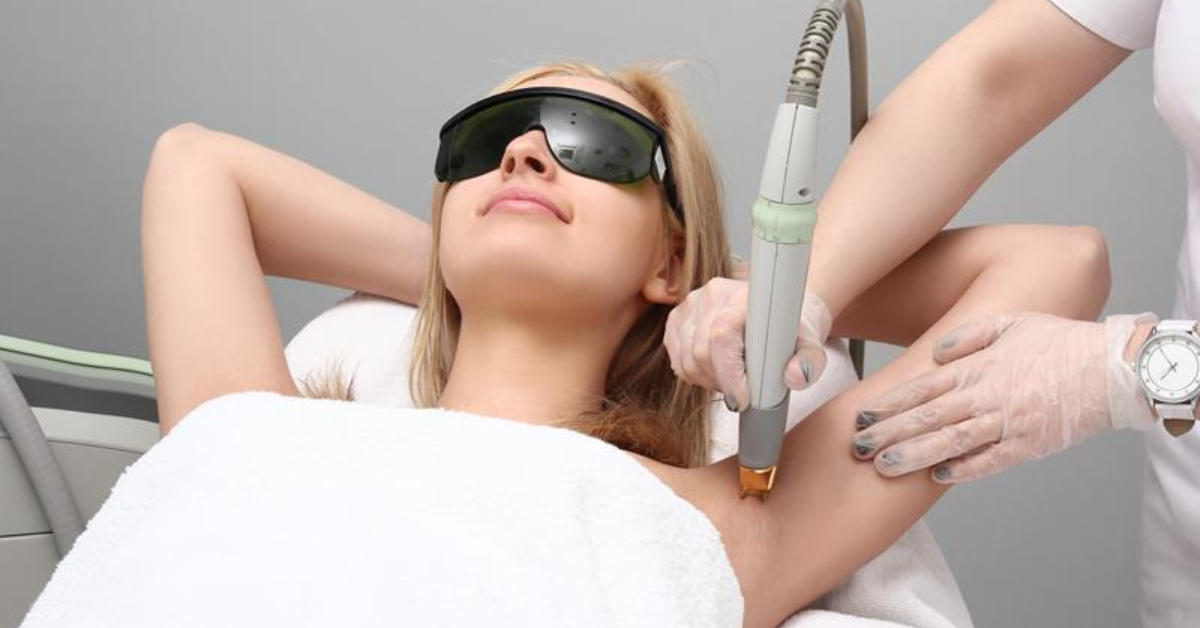 Women laying in white body cover wearing black  laser eye protection glasses while the other woman doing her laser for underarms